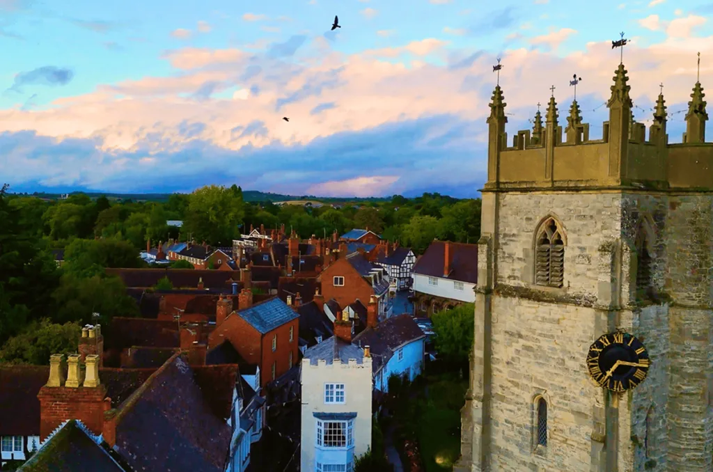 Charming English Towns in the West Midlands An elevated view of Alcester showing the historic stone tower and clock face of St Nicholas Church, overlooking traditional red-brick and timber-framed rooftops with green countryside hills under a sunset sky.