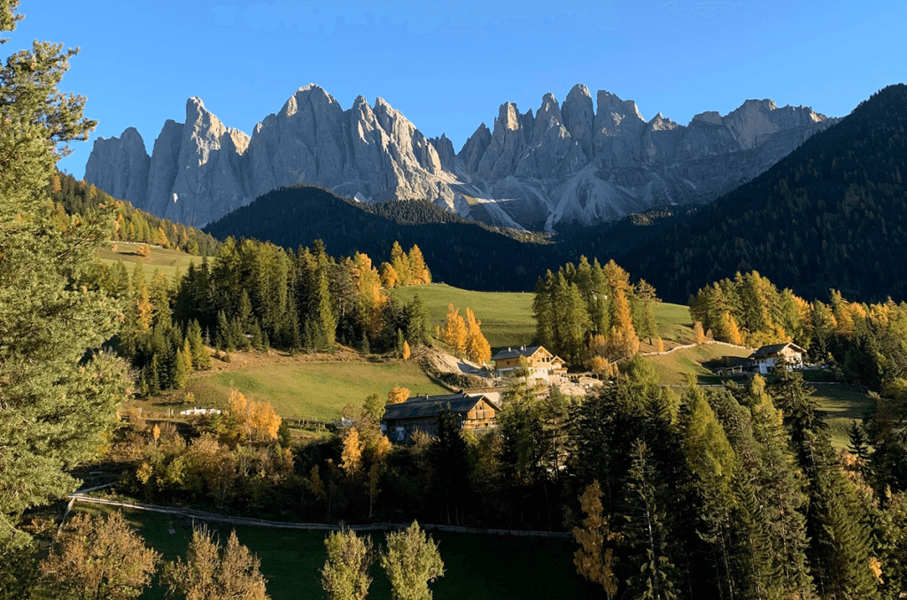 Circle Itinerary for the Dolomites Dramatic jagged peaks of the Odle Mountains in the Italian Dolomites, rising above a sunlit valley with autumn-colored trees and traditional alpine houses.