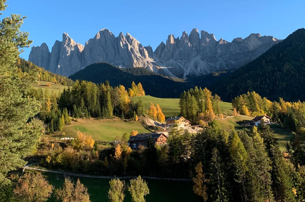 Dramatic jagged peaks of the Odle Mountains in the Italian Dolomites, rising above a sunlit valley with autumn-colored trees and traditional alpine houses.