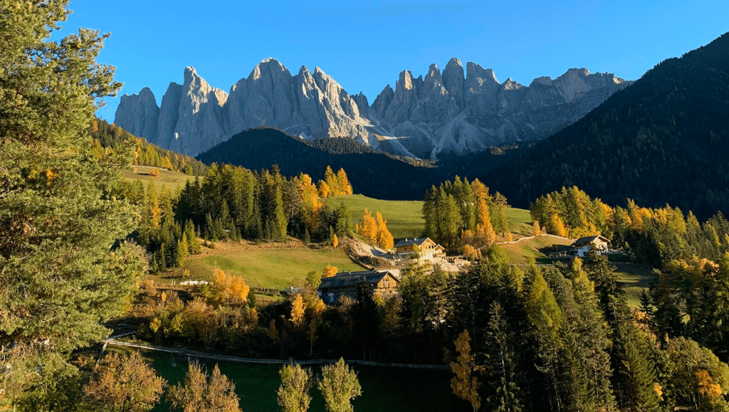 Dramatic jagged peaks of the Odle Mountains in the Italian Dolomites, rising above a sunlit valley with autumn-colored trees and traditional alpine houses.
