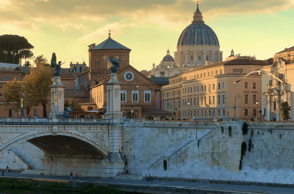 What to See in Rome Panoramic view of Rome at sunset with St. Peter's Basilica dome and a historic bridge over the Tiber River in the foreground.