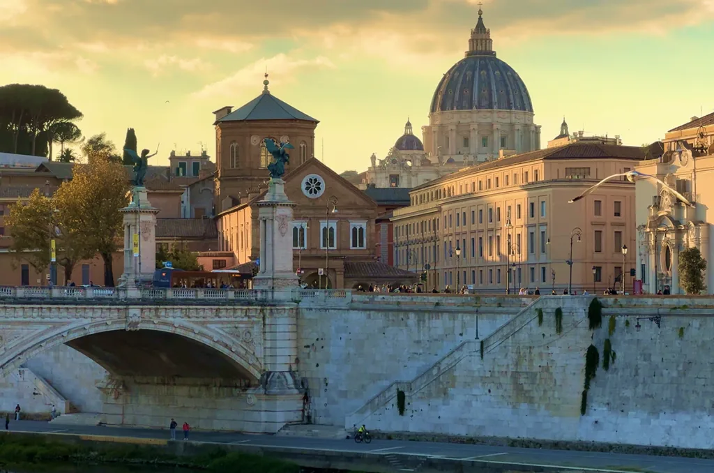 Panoramic view of Rome at sunset with St. Peter's Basilica dome and a historic bridge over the Tiber River in the foreground.