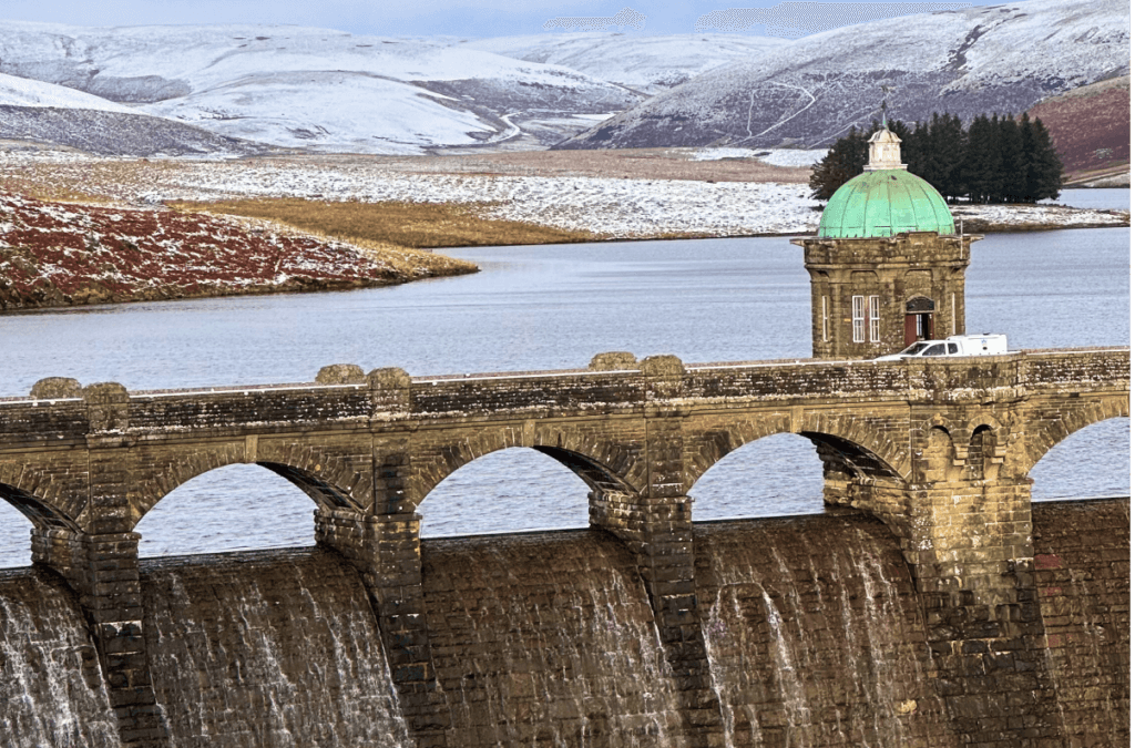 Winter Weekend in Wales A wide view of the Pen y Garreg Dam in the Elan Valley, Wales, during winter, featuring the stone dam wall with water cascading over it and a green-domed tower overlooking the reservoir and snow-dusted hills.