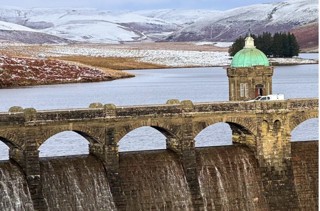 Winter Weekend in Wales A wide view of the Pen y Garreg Dam in the Elan Valley, Wales, during winter, featuring the stone dam wall with water cascading over it and a green-domed tower overlooking the reservoir and snow-dusted hills.