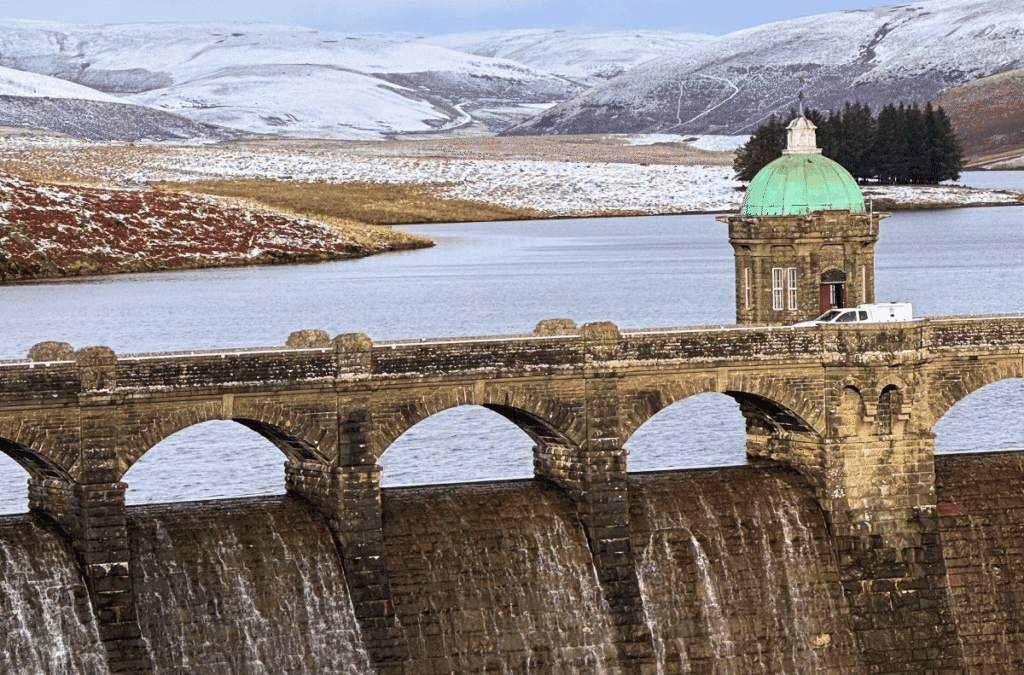 A wide view of the Pen y Garreg Dam in the Elan Valley, Wales, during winter, featuring the stone dam wall with water cascading over it and a green-domed tower overlooking the reservoir and snow-dusted hills.