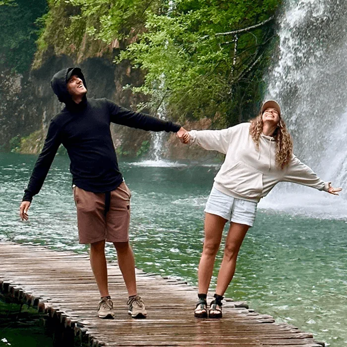 A man and a woman holding hands and smiling on a wooden walkway in front of a large waterfall at Plitvice Lakes National Park.