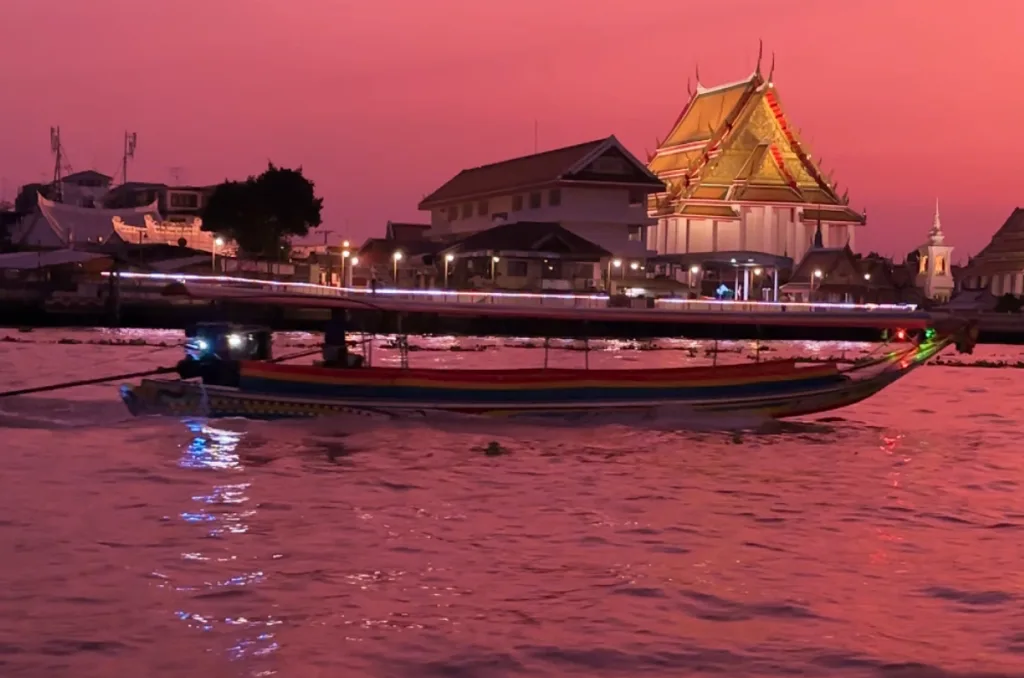 A traditional Thai long-tail boat cruising on the Chao Phraya River in Bangkok during a vivid pink and orange sunset, with a golden-roofed Buddhist temple illuminated on the riverbank.