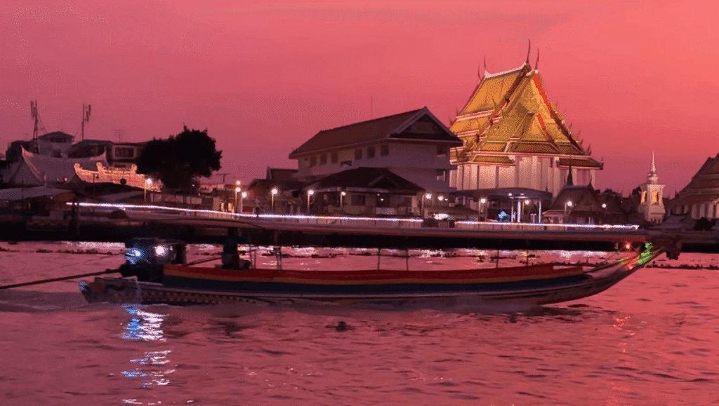 A traditional Thai long-tail boat cruising on the Chao Phraya River in Bangkok during a vivid pink and orange sunset, with a golden-roofed Buddhist temple illuminated on the riverbank.