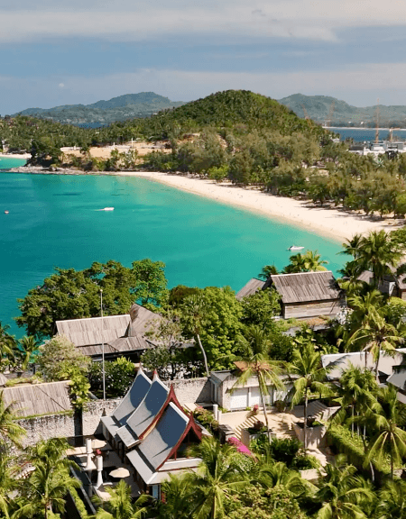 A sweeping high-angle view of Pansea Beach in Phuket, Thailand, Asia, featuring luxury villa rooftops with traditional Thai architecture nestled in lush palm trees overlooking a white sand crescent and turquoise ocean.