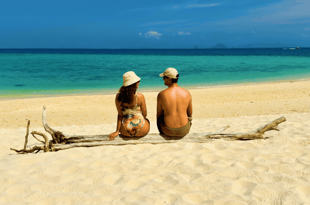 A couple seen from behind, sitting on a large piece of sun-bleached driftwood on a powdery white sand beach in Phuket, Thailand, looking out at vibrant turquoise water and distant limestone karst islands.