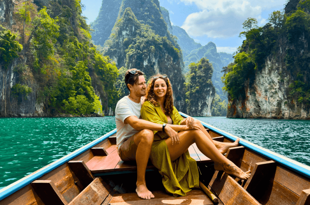 A couple sitting on the wooden bow of a traditional Thai long-tail boat on the emerald green water of Cheow Lan Lake in Khao Sok National Park, surrounded by massive limestone cliffs and lush jungle.