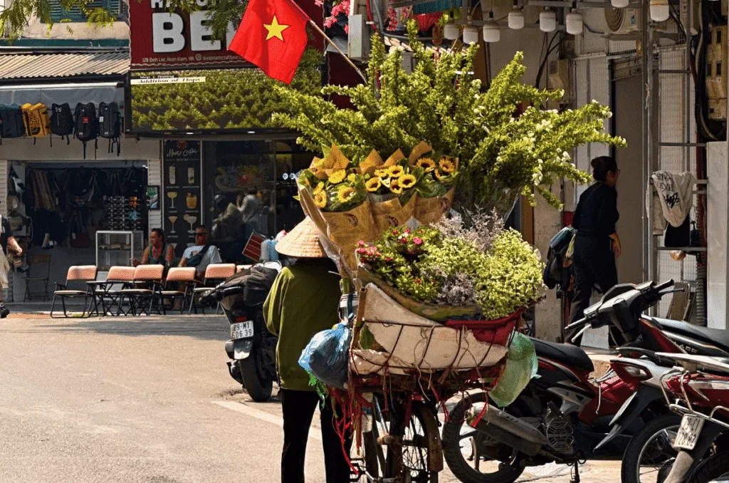 A local Vietnamese street vendor wearing a traditional conical hat (nón lá) pushing a bicycle laden with fresh yellow sunflowers and green bouquets down a sunny Hanoi Old Quarter street with parked motorbikes.